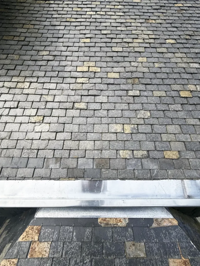 Gros plan sur une couverture en ardoise naturelle panachée, fixée avec des crochets, et sur un chéneau neuf en zinc.