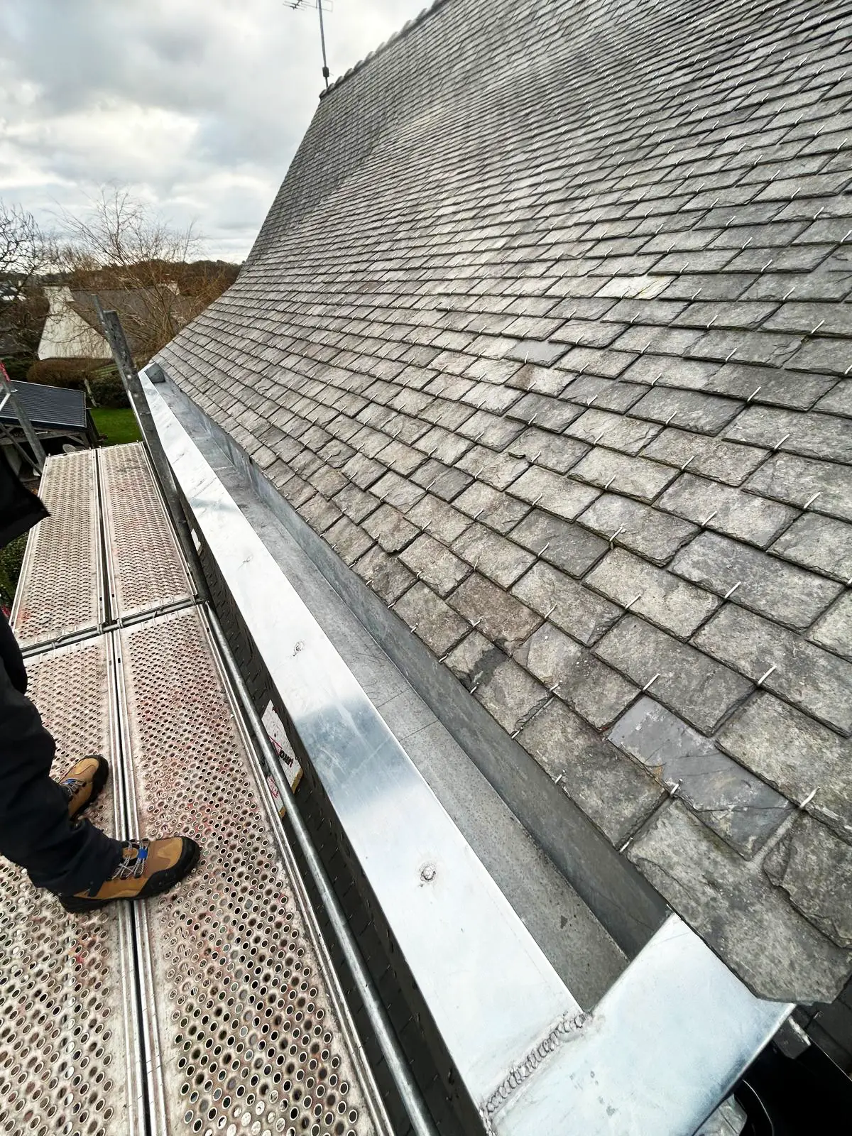 Pose d'un chéneau en zinc sur mesure le long d'une toiture en ardoise, vu depuis l'échafaudage.